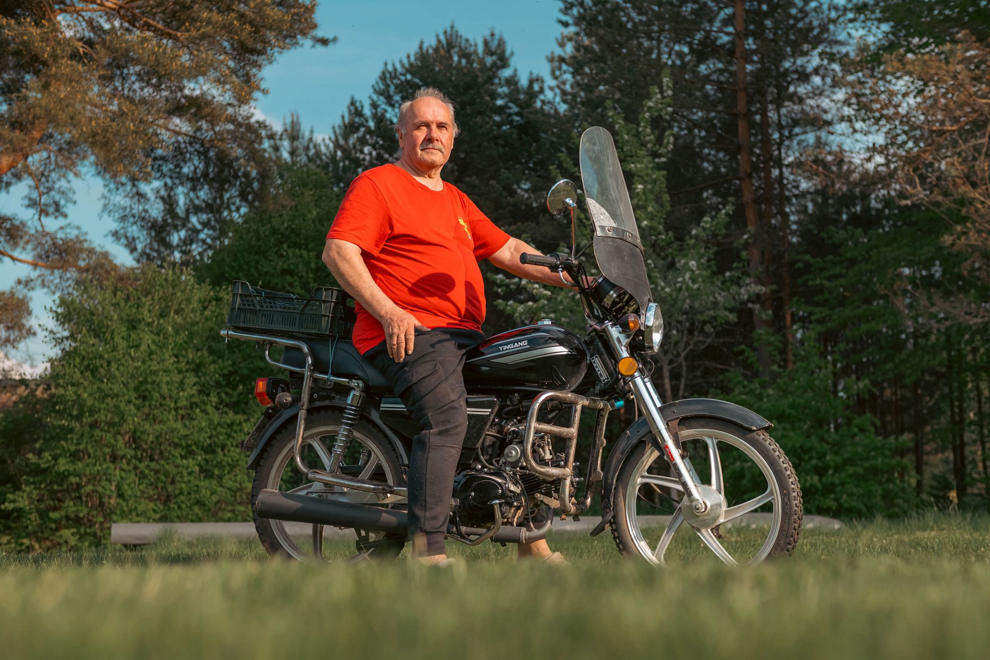 Aleksandrs sēž uz motocikla pagalmā.