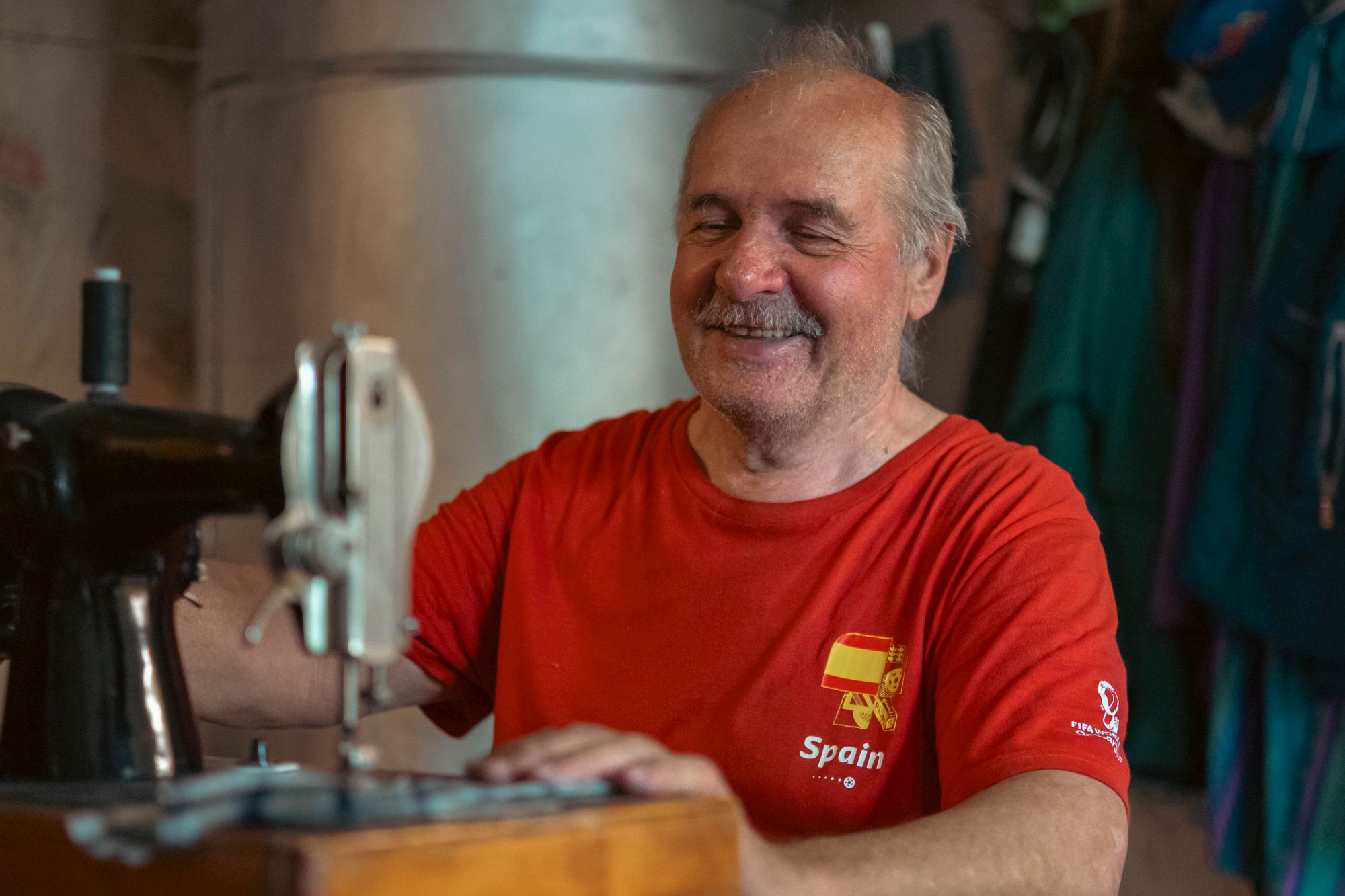 Aleksandrs sēž blakus šujmašīnai un smaida.