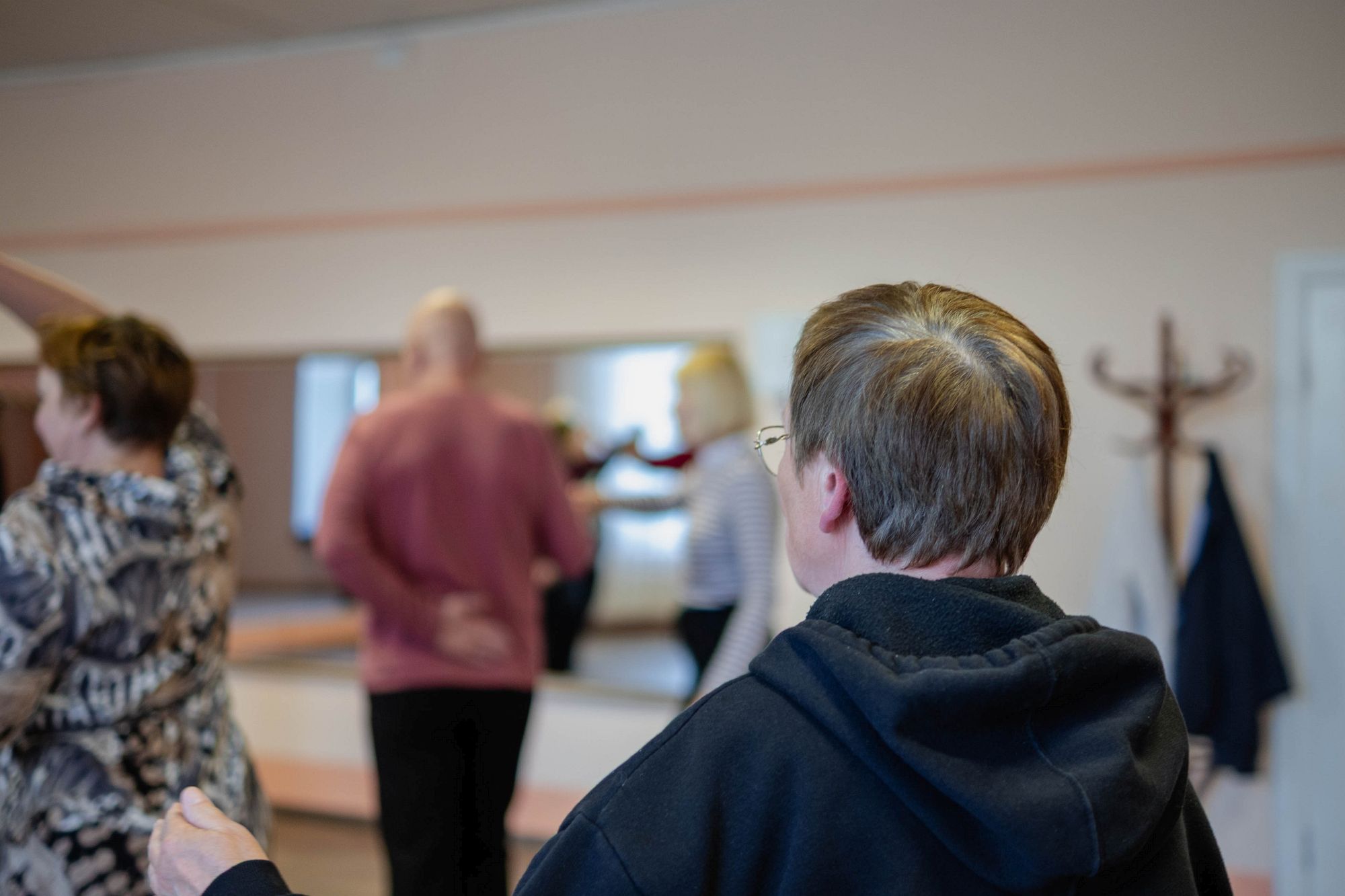 Jeļena skatās uz nodarbības dalībniekiem; cilvēki dejo spoguļa priekšā, viņa vēro no malas.