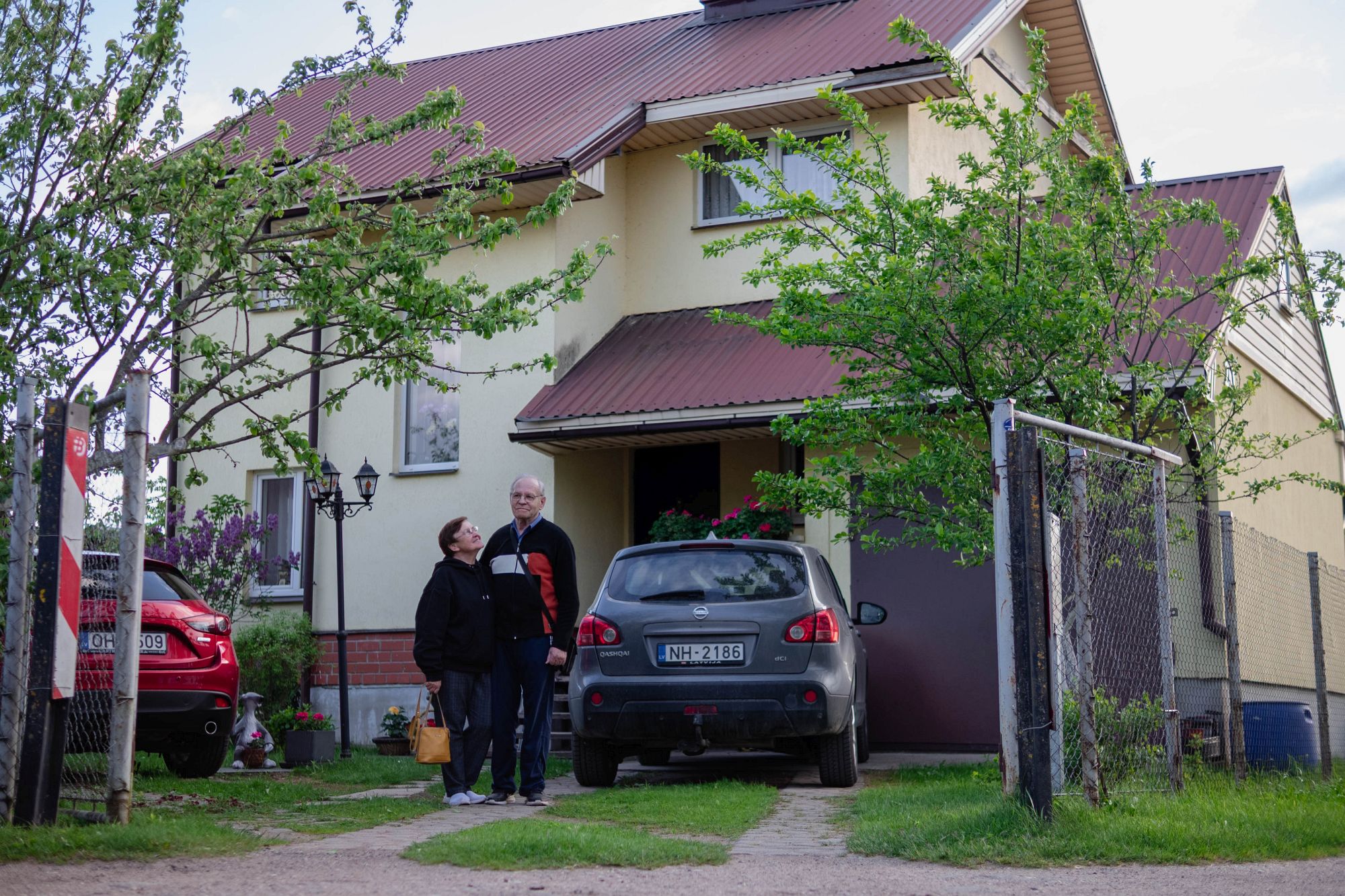 Jeļena un viņas vīrs stāv pagalmā pie mājas; māja, automašīnas, koki, atvērti vārti.