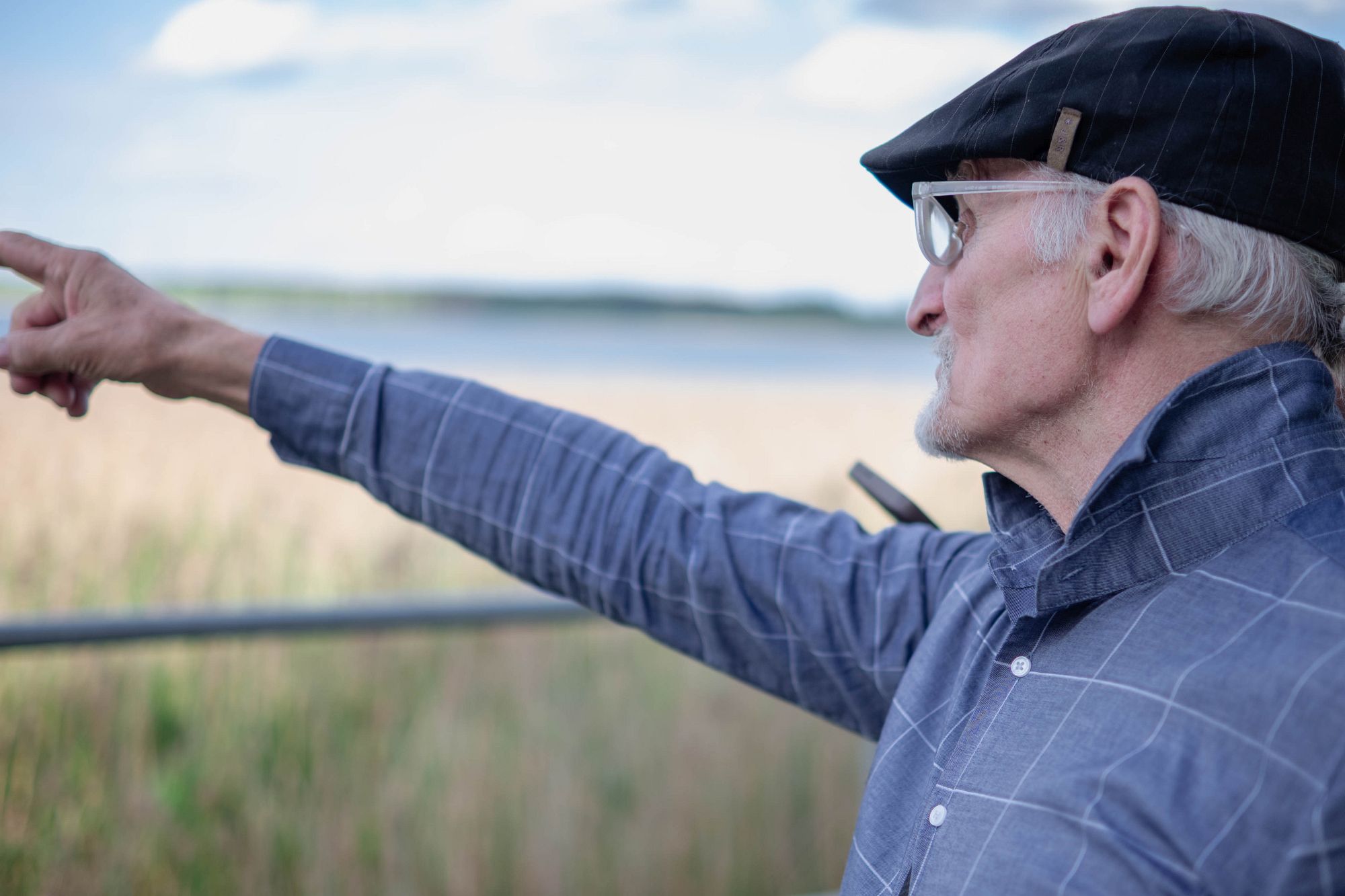 Jurijs profilā ar roku rāda tālumā. Viņš stāv pie ūdens, brillēs un cepurē; žests ir svarīgāks par seju — viņš skaidro, kur bijusi sporta distance.