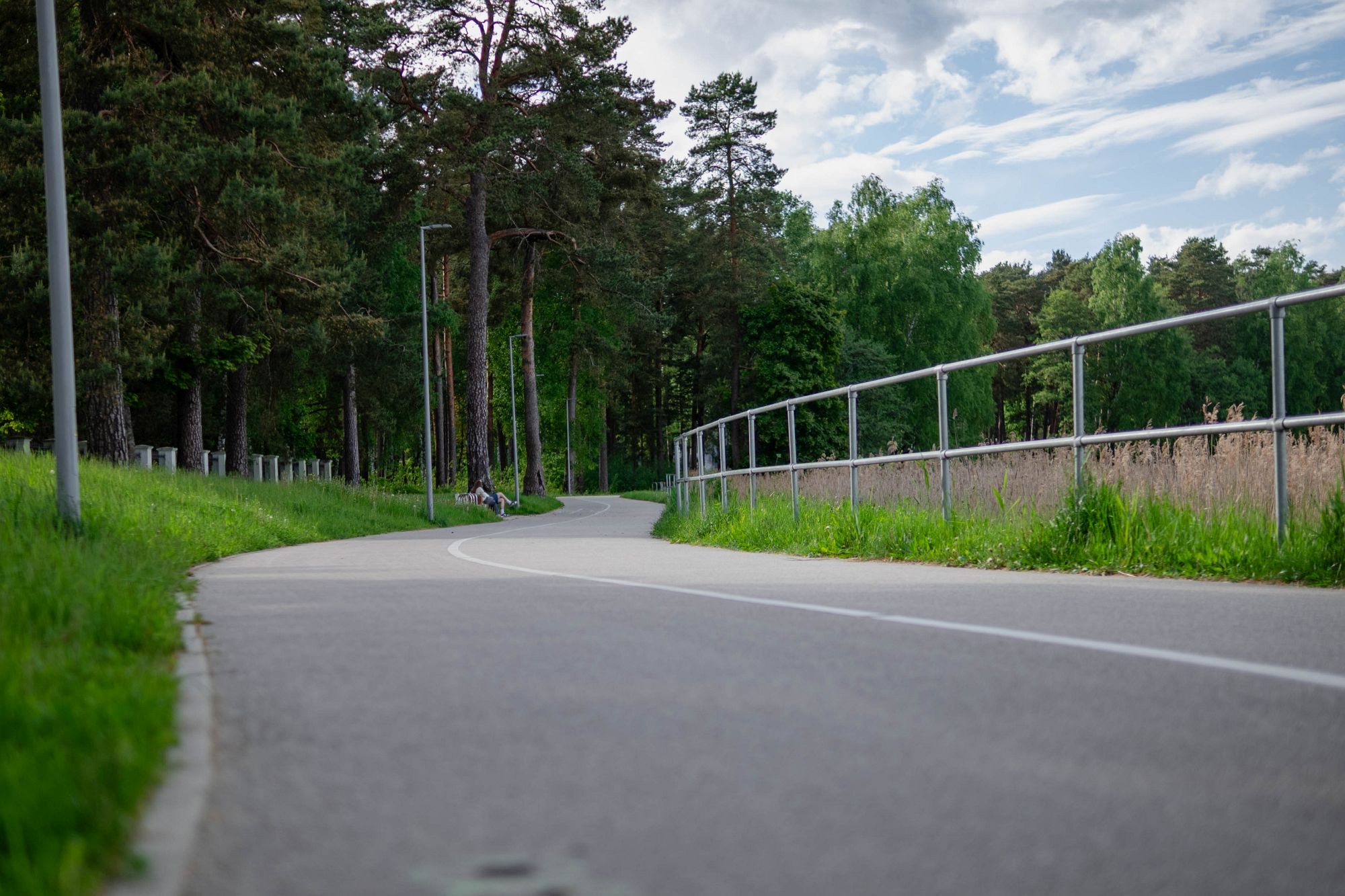 Tukšs celiņš zaļajā zonā pie ūdens. Ceļa līnija ved skatienu uz priekšu, pa labi metāla margas, apkārt koki un zāle.