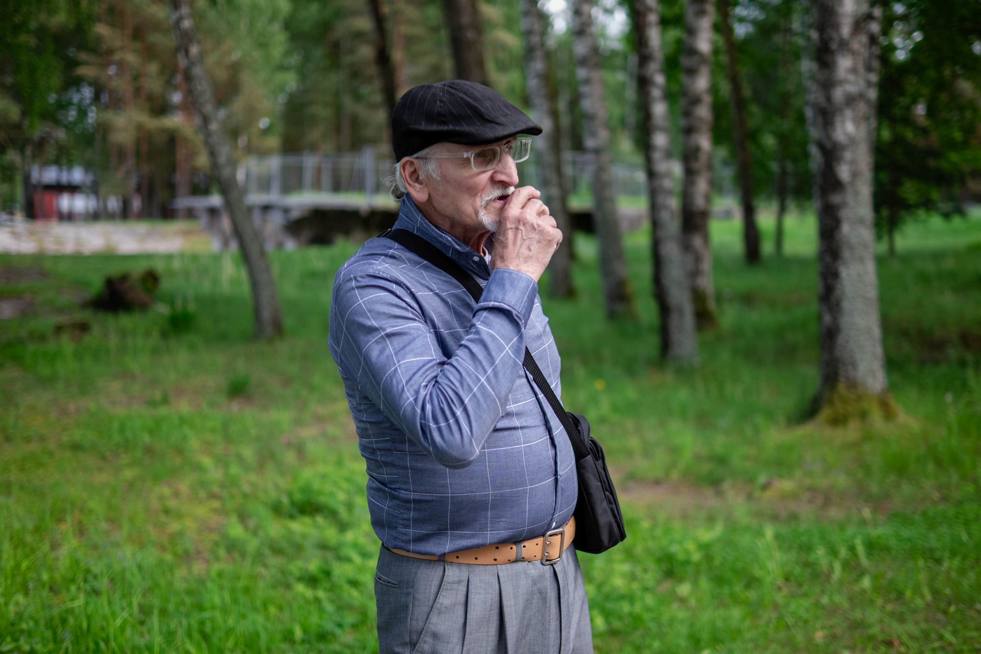 Jurijs koku vidū zaļajā zonā, roka pie sejas, it kā uz brīdi apstādamies sarunā. Fonā tiltiņš un ūdens.