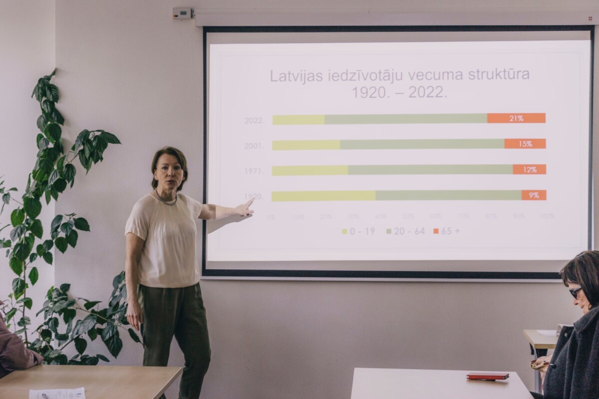 Tatjana Azamatova tikšanās ar Naujenes pagasta iedzīvotājiem rāda slaidu, kurā parādīts, kā pēdējo 100 gadu laikā Latvijā ir mainījusies iedzīvotāju vecuma attiecība. 2025. gada 12. maijs, Naujenes kultūras centrs. Foto: Jevgēņijs Ratkovs
